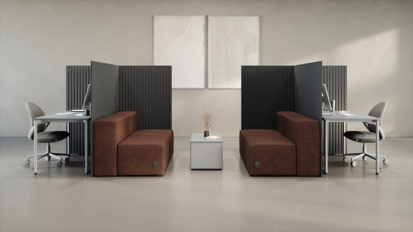 Minimalist office space with two separate workstations, partitioned by dark panels. Each station has a chair, desk, and lamp. A central seating area features brown couches and a small table
