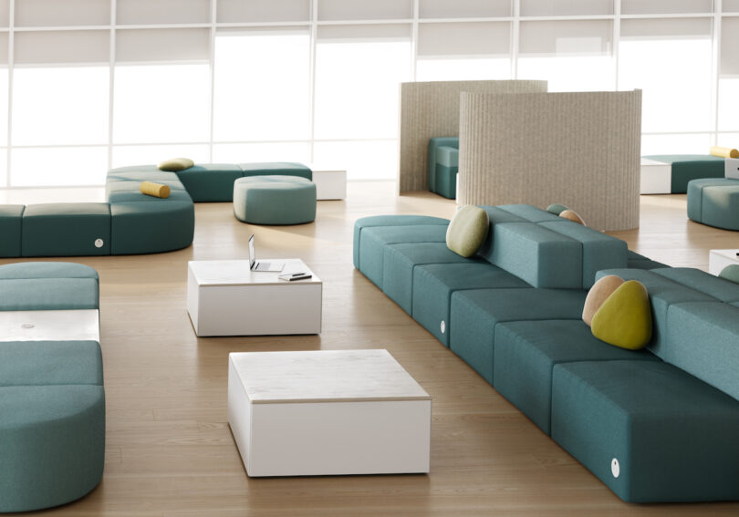 Spacious modern lounge with teal modular seating and white tables, sunlight streaming through tall windows in the background