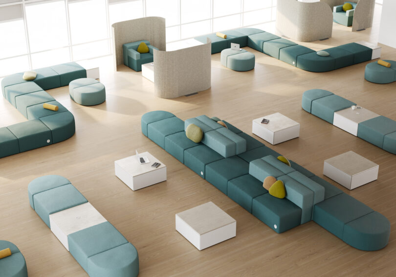 A modern, open lounge area with modular teal seating, white tables, and cushions on wood flooring, lit by natural light from large windows