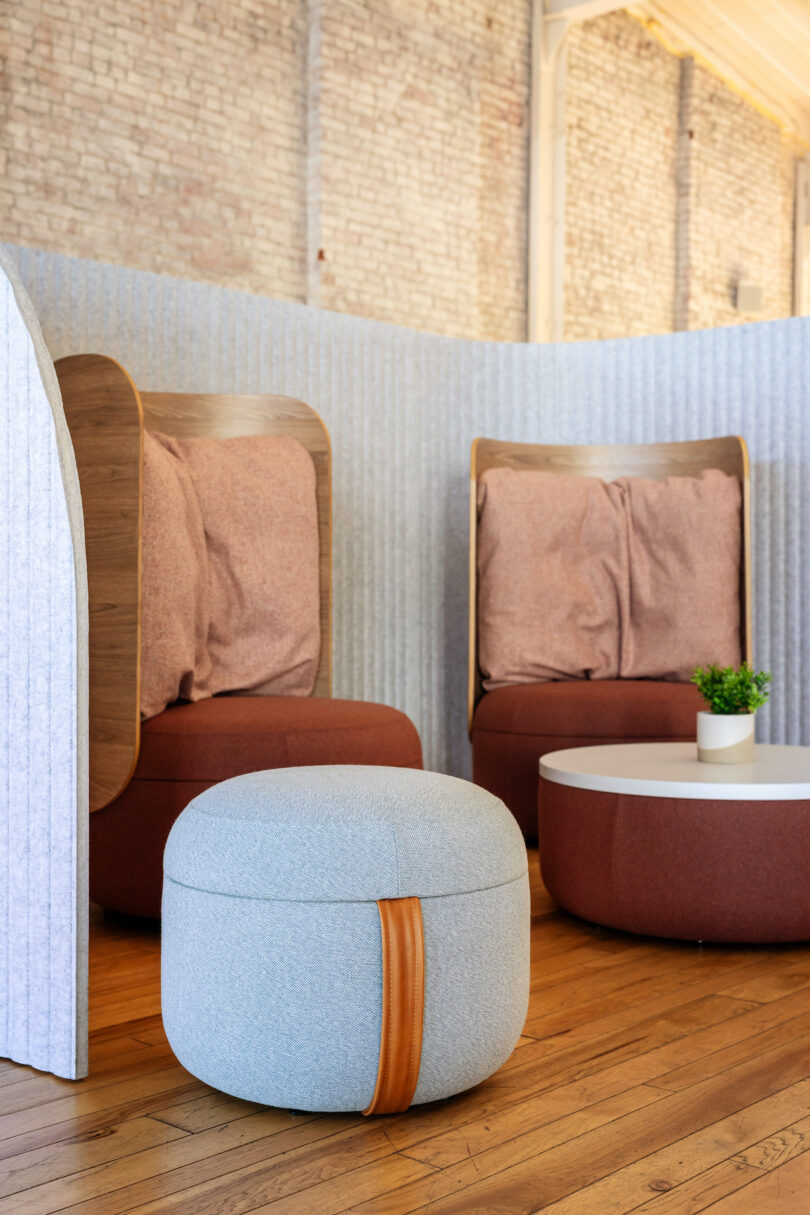 Modern lounge area with two cushioned chairs, a round ottoman, and a small coffee table with a plant, set against a brick wall and partition