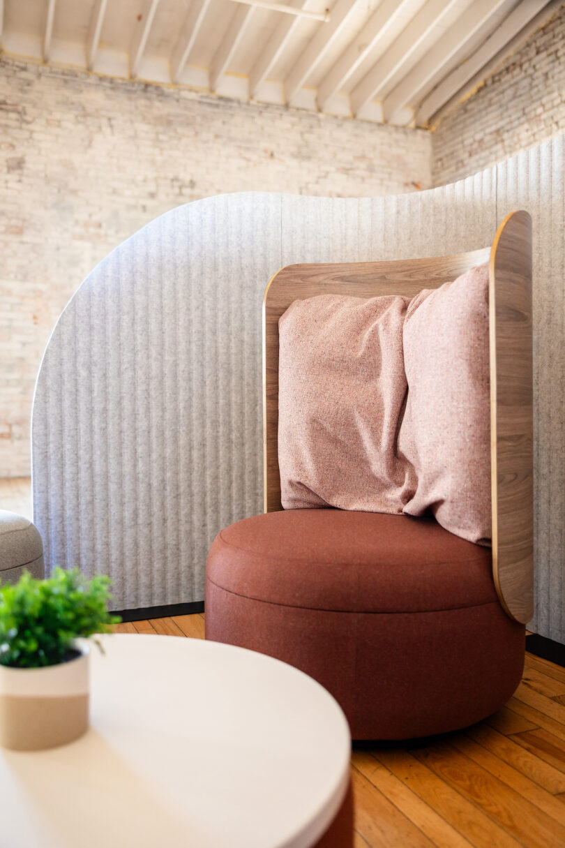Modern lounge chair with a curved wooden back and pink cushions, set on a wooden floor. A white table with a small green plant is in the foreground