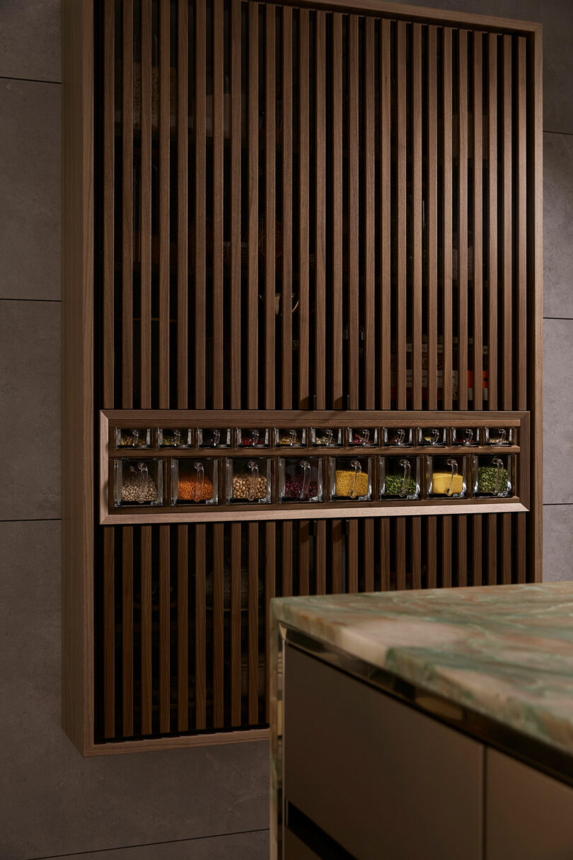 A wooden cabinet with vertical slats and a row of visible spice jars, viewed from a kitchen. A countertop with a green surface is in the foreground.