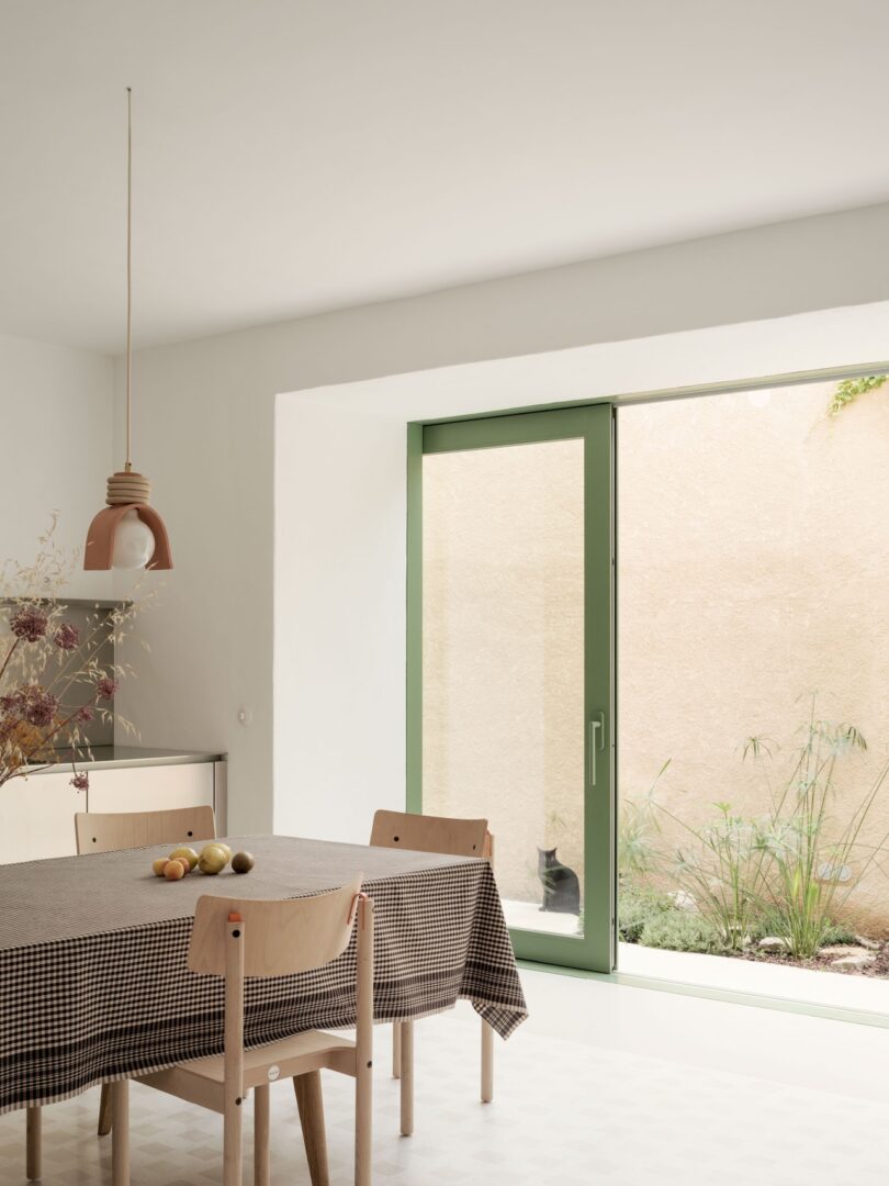 Minimalist dining area with a table and chairs, covered with a checkered cloth. Pendant light above. Large window with green trim shows a plant and a cat outside.
