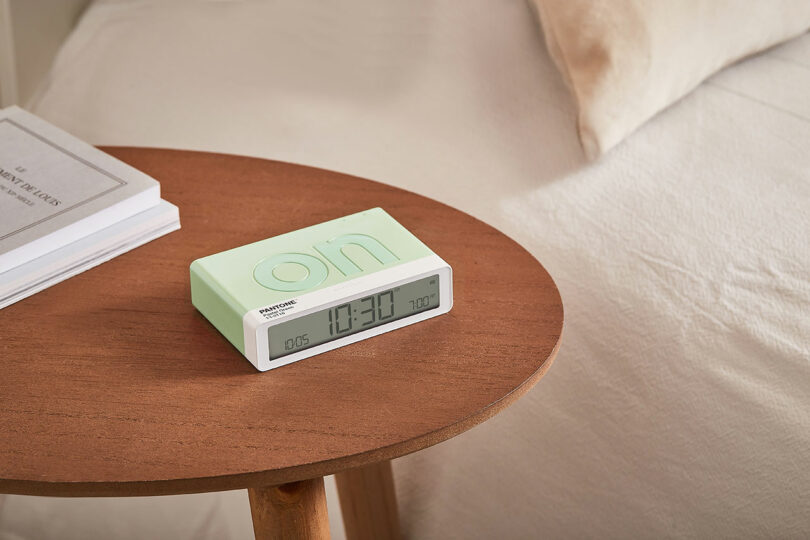 A digital alarm clock displaying 10:30 is placed on a round wooden bedside table, next to a bed with beige and white linens.