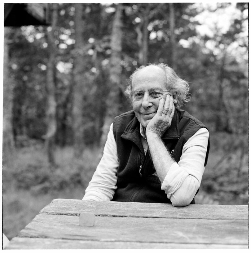 A person seated at a wooden table outdoors, resting their head on their hand, smiling with an Alex Matisse aura. Trees fill the background. Black and white photo.
