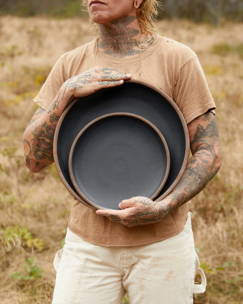 A person with tattoos, reminiscent of Alex Matisse's artistry, holds two black circular plates of different sizes in an outdoor setting.