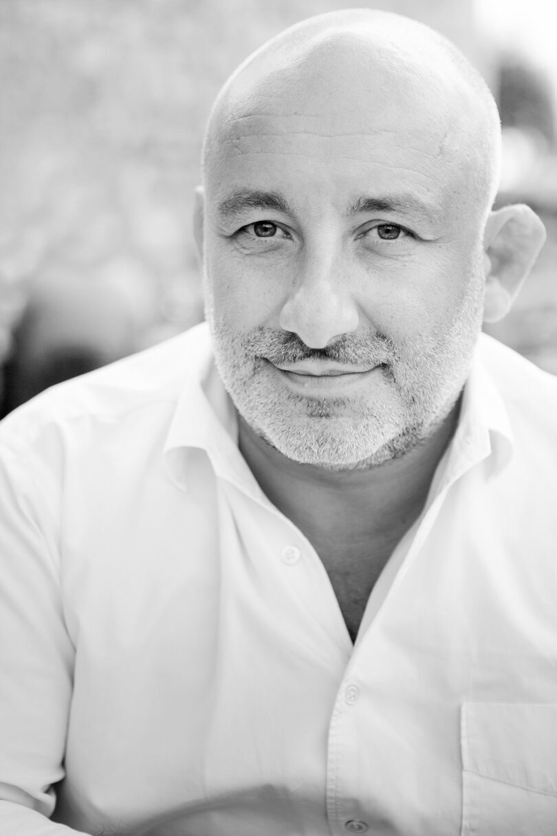 A bald man with a salt-and-pepper beard wearing a white shirt looks directly at the camera in this black-and-white photo.