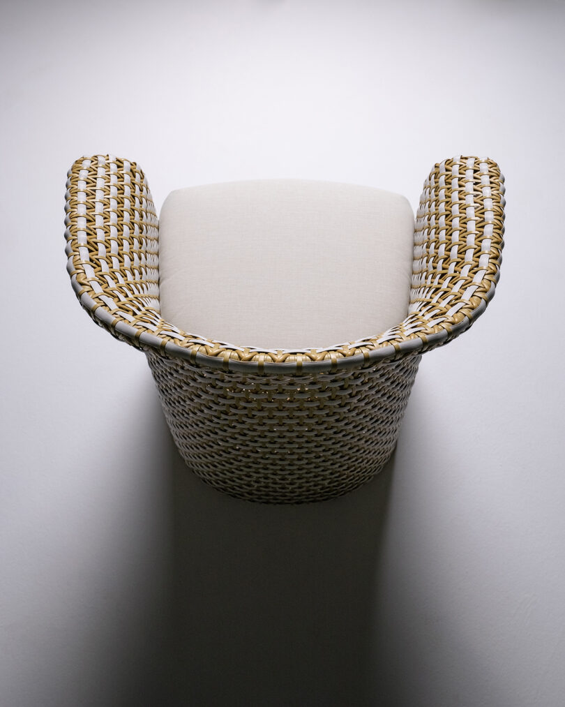 Overhead image of a wicker chair with a beige cushion seat, featuring a curved backrest and armrests, set against a plain, light background.
