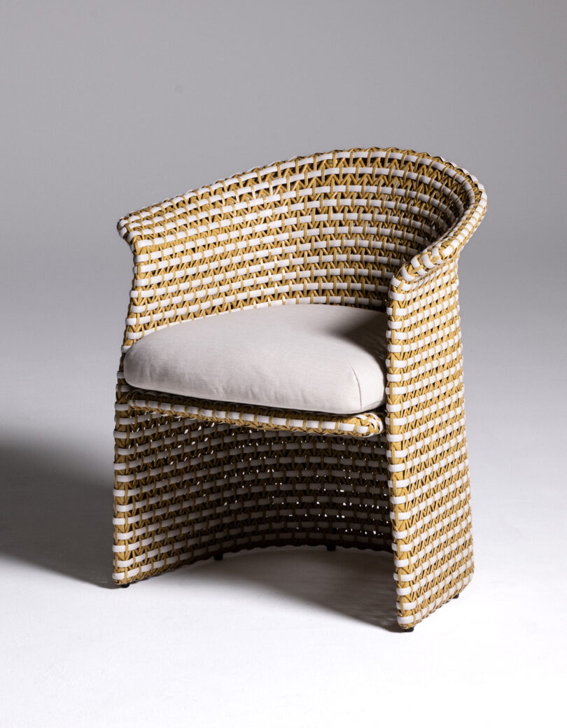 A wicker chair with a beige cushion seat, featuring a curved backrest and armrests, set against a plain, light background.