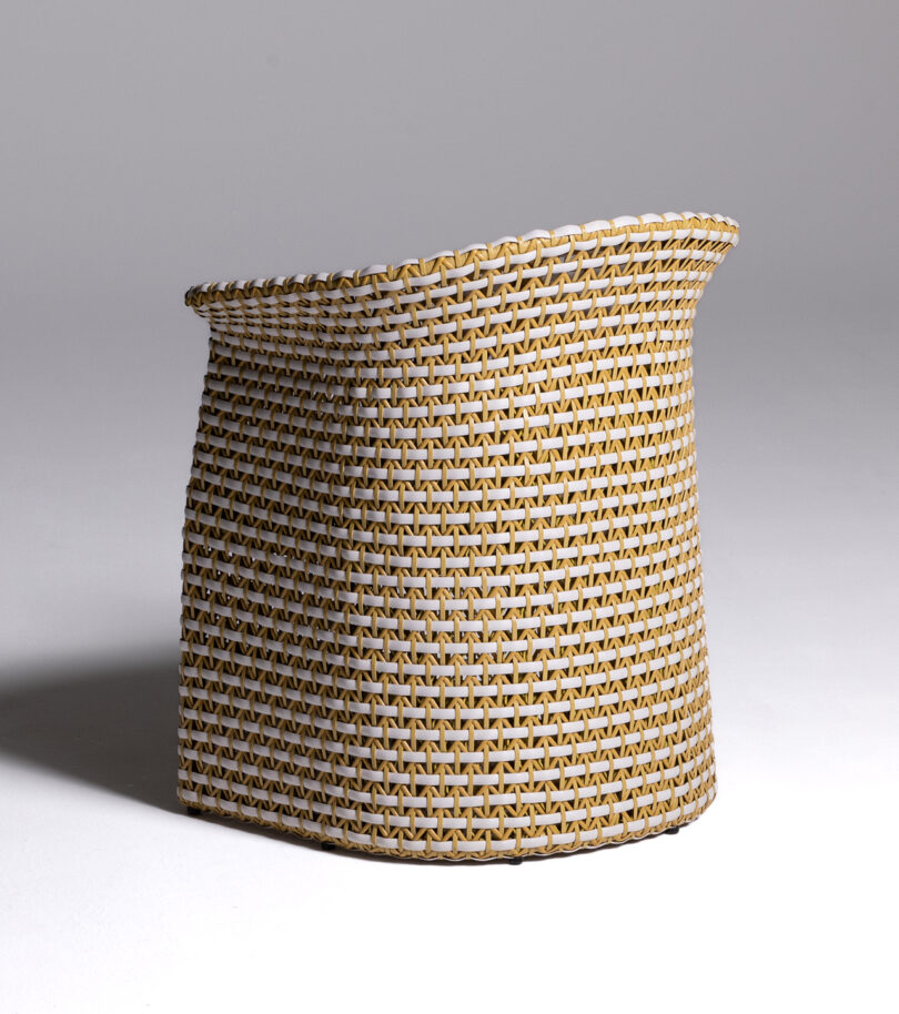 Back of a wicker chair with a beige cushion seat, featuring a curved backrest and armrests, set against a plain, light background.