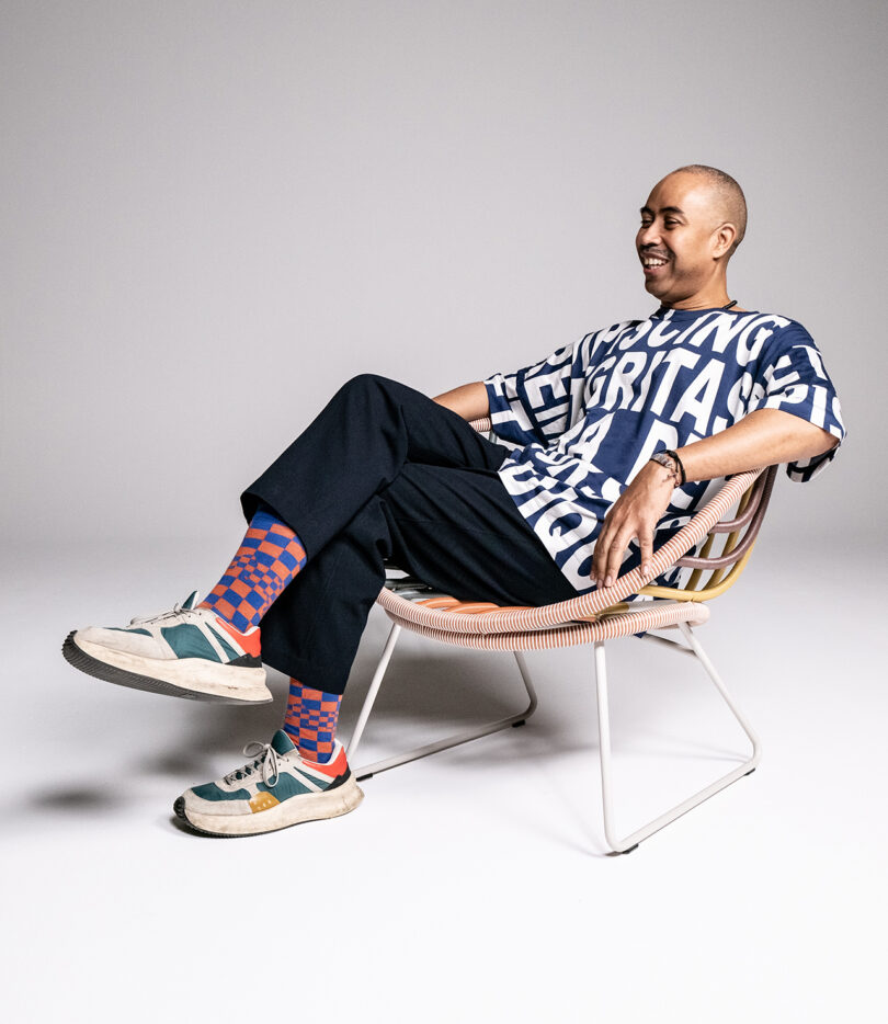 A person wearing a patterned shirt, black pants, and colorful socks with sneakers sits casually in a modern chair against a plain background.