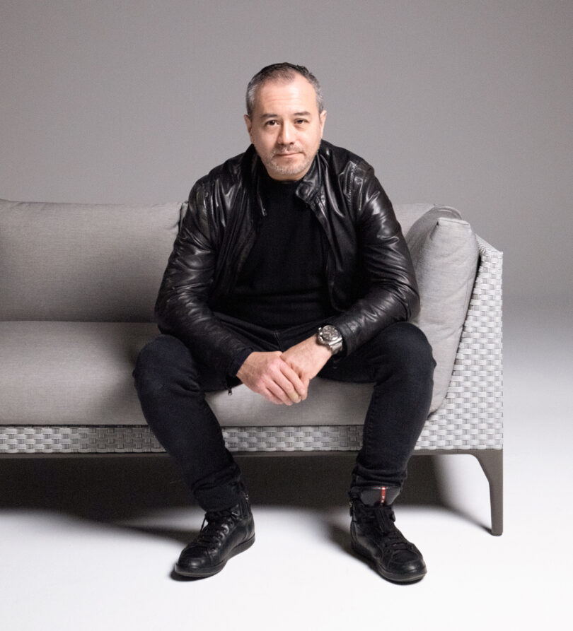 A man with short hair and a beard sits on a gray couch wearing a black leather jacket, black pants, and black shoes, with his hands clasped and looking towards the camera.