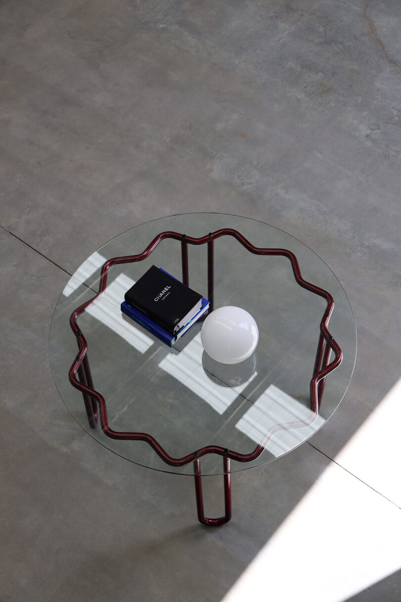 The Yonder Table features a wavy red metal frame and a sleek glass top, adorned with a stack of books and a white spherical object. This Tubulaa masterpiece rests on the concrete floor, adding an artistic touch to any space.
