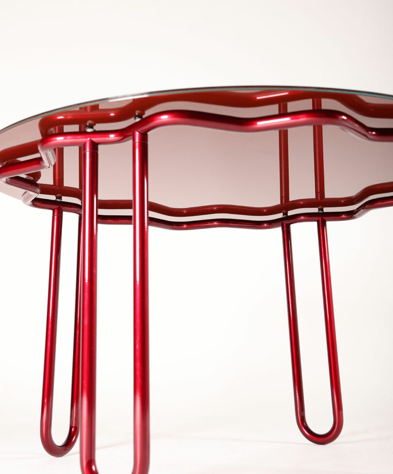 The Yonder Table features a striking red, curved metal frame and a wavy-edged glass top, presented elegantly on a white background.