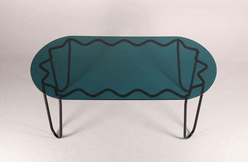 The Yonder Table Tubulaa is an oval glass masterpiece with a green tint, adorned with black wavy lines beneath the surface. It stands gracefully on slender, curved black legs, offering a unique blend of elegance and modern design.