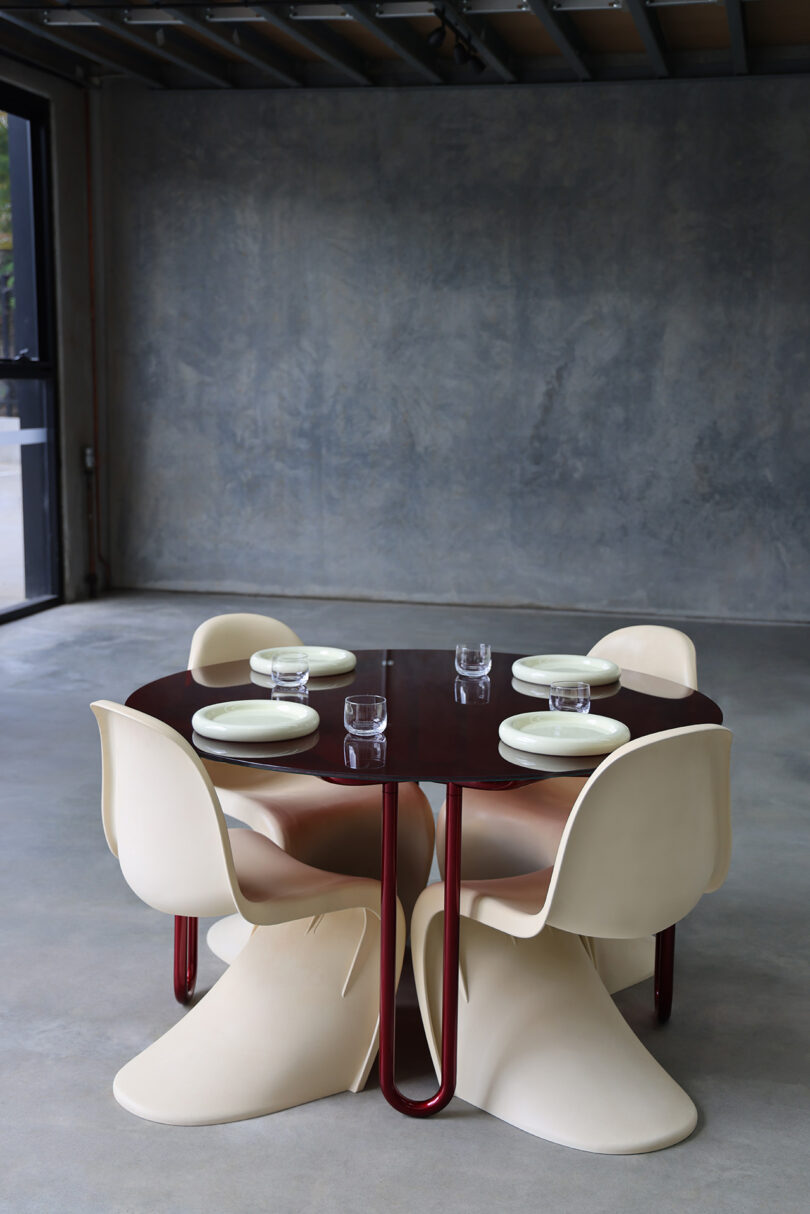 A modern dining setup featuring the Yonder Table, a round dark wooden masterpiece, accompanied by Tubulaa's uniquely shaped cream-colored chairs. The minimalist room is perfectly arranged with four place settings of empty white plates and gleaming glasses.