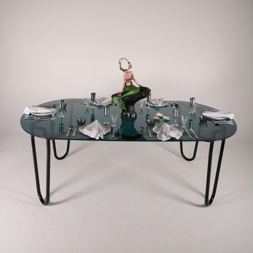 The Yonder Table Tubulaa dining set, designed for four, showcases silverware, plates, and napkins alongside a striking central sculpture of a seated figure in a green dress.