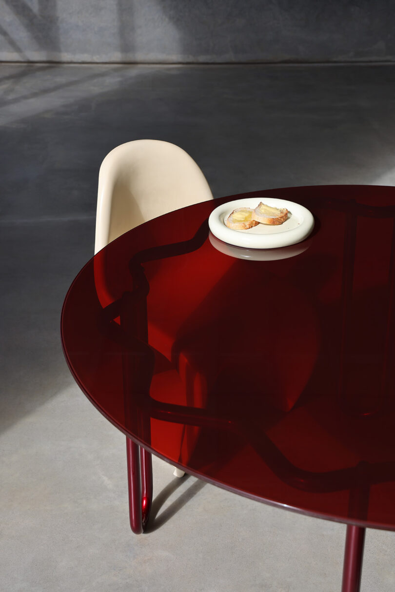 A round, dark red Yonder Table with thin legs holds a white plate with sliced bread. A beige, sculptural chair is situated behind the table.