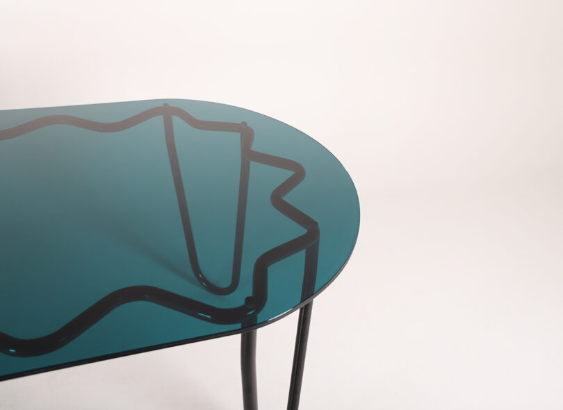 The Yonder Table Tubulaa boasts a blue-tinted glass top with sleek black metal legs, accented by an elegant wavy black line pattern beneath the surface.