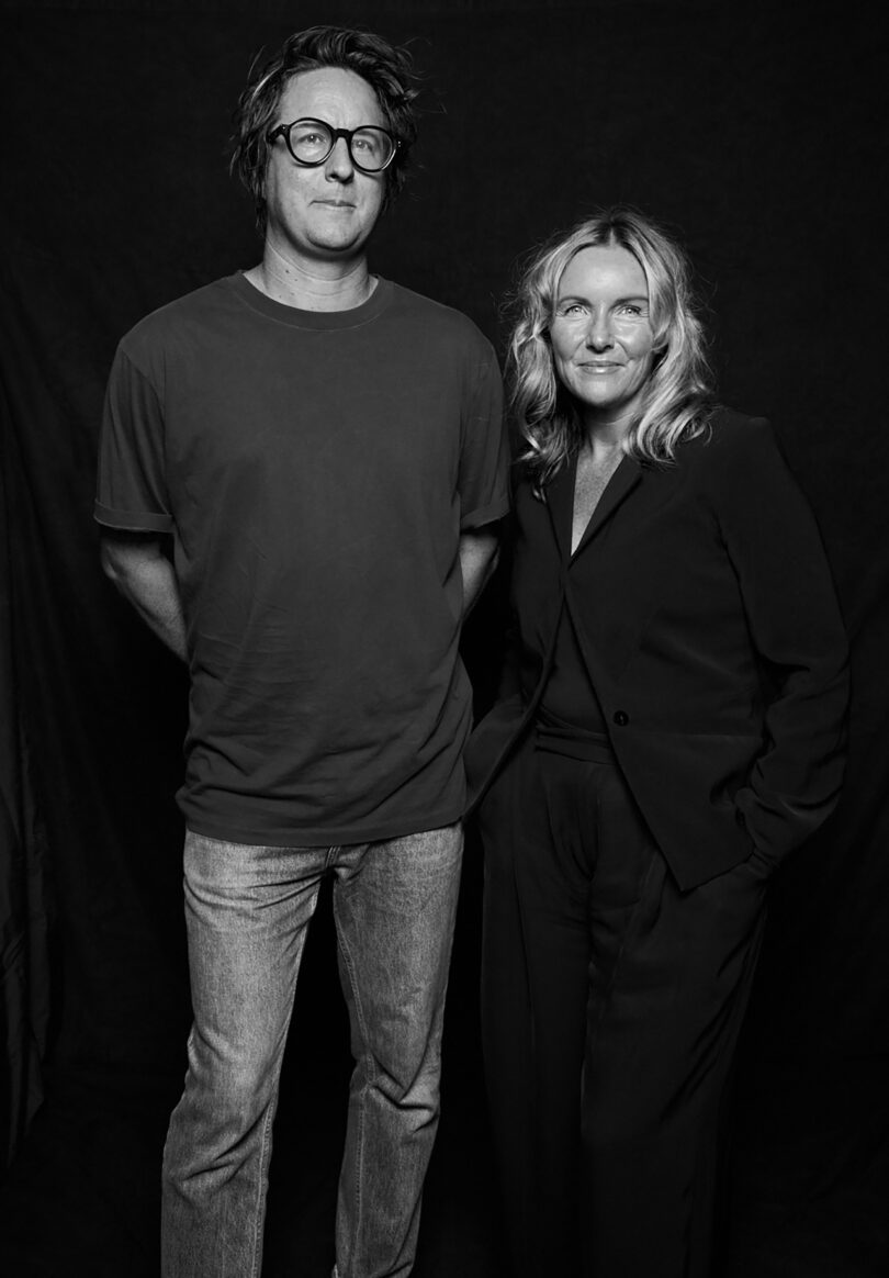 A man in a t-shirt and jeans stands next to a woman in a suit, both facing forward, against the dark backdrop of Yonder.