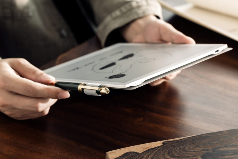 Person holding a tablet displaying a sketch, with a pen resting on it, over a wooden table.
