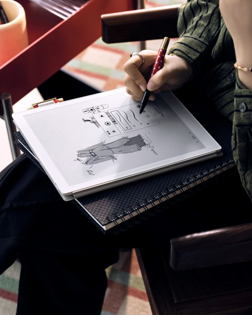 Person sketching clothing designs on a tablet using a stylus, with a notebook underneath.
