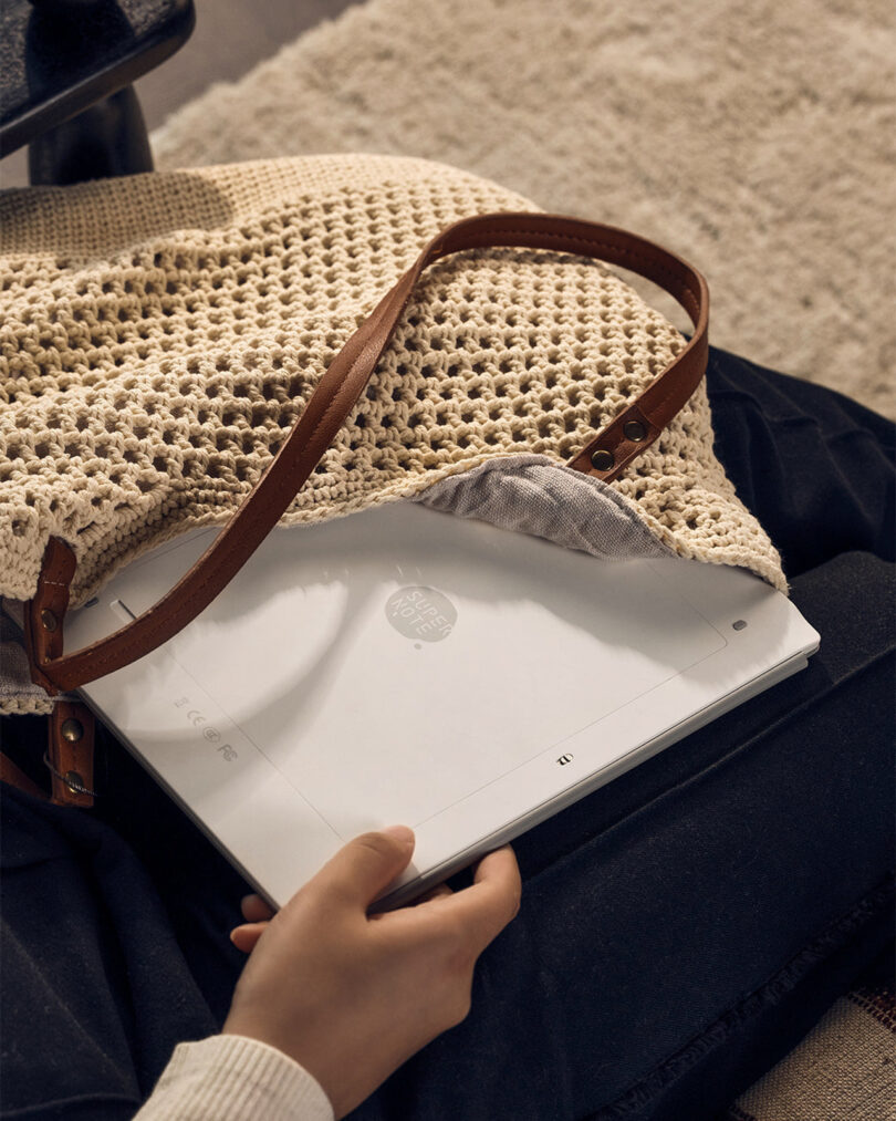A person holding a white electronic device, partially pulled out from a beige, knitted bag with a brown leather strap.