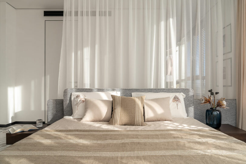 Modern bedroom with a neatly made bed, light beige bedding, and decorative pillows. Sheer curtains hang above, with sunlight streaming in. A book and vase with dried flowers are on the bedside tables.