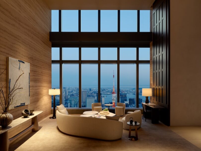Luxurious living room with modern furniture, large windows revealing a cityscape including a prominent tower, illuminated by soft interior lighting.