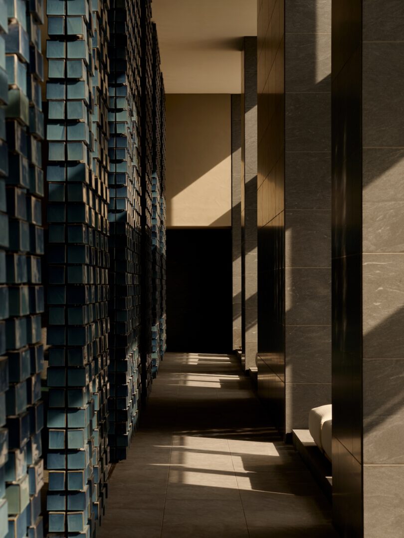 A narrow hallway is lined with gray tile and textured blue wall panels, with sunlight casting geometric shadows on the floor.