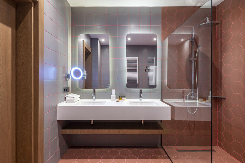 Nestled in a style reminiscent of Lopota Lake Resort, the modern bathroom features two sinks, two mirrors, and a glass shower enclosure. The walls and floors are adorned with hexagonal red tiles, while a lit vanity mirror adds elegance on the left.