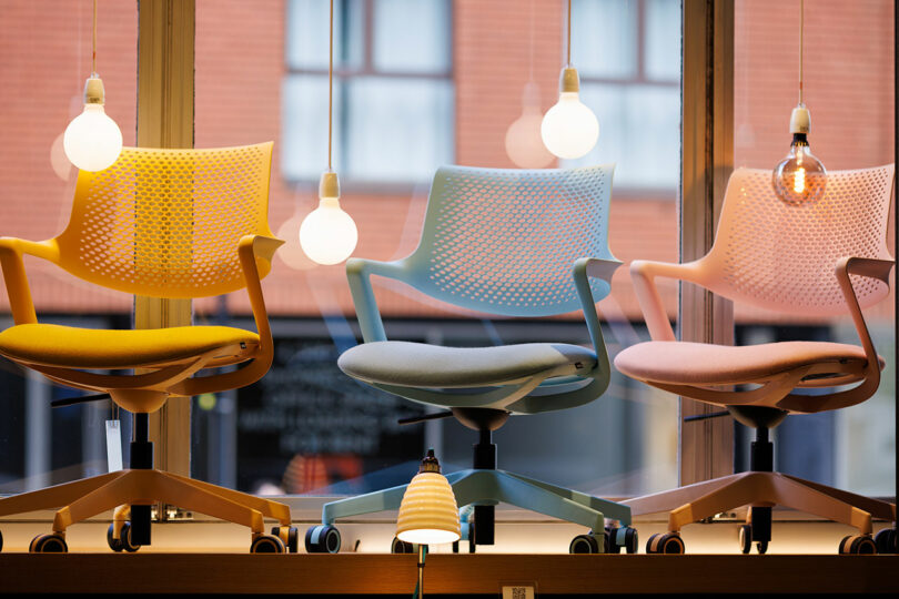 Three colorful office chairs with perforated backs are lined up by a window, their sleek design hinting at the innovation celebrated at the CDW 2025 Product Awards. Hanging light bulbs illuminate the scene, casting a warm glow on these award-worthy pieces.