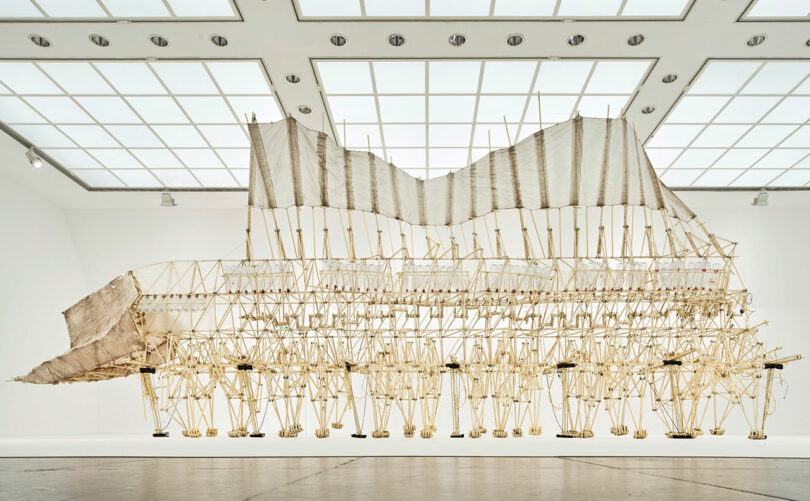 Large kinetic sculpture resembling a skeletal creature with numerous legs, set in a bright, spacious gallery with a grid-patterned ceiling.