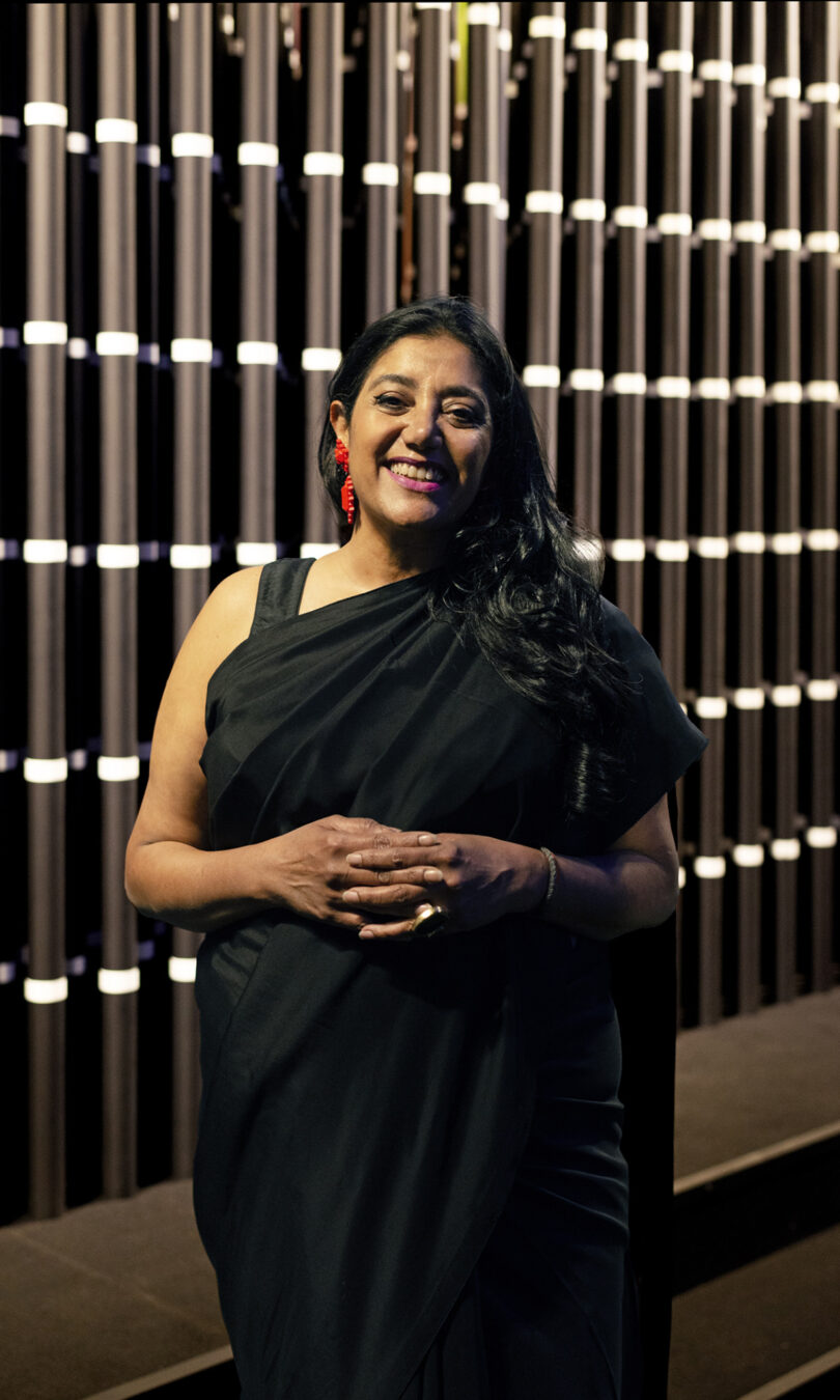 A person in a black outfit stands smiling in front of a patterned background, with folded hands.