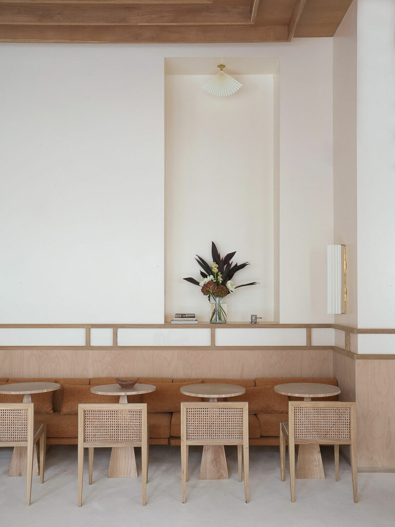 The minimalist cafe interior features Policronica-inspired light wood furniture, a brown sofa, small round tables, and a flower arrangement in a vase on a shelf.