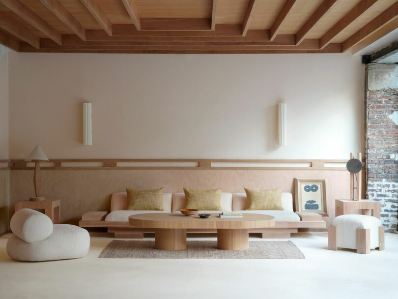 In a minimalist living room, the wooden ceiling and beige walls frame a Policronica-inspired low wooden sofa. Two round chairs and a large coffee table complement modern wall lights, while a textured rug adds subtle depth to the space.