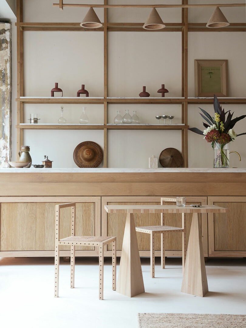 The minimalist kitchen features Policronica wooden shelves, a table, and chairs. Shelves display vases, glasses, and a framed picture. A vase filled with vibrant flowers sits on the countertop, while stylish pendant lights hang above.