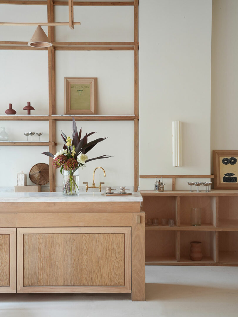 A minimalist kitchen features wooden cabinets, a white countertop, and open shelving. A vase with flowers sits on the counter, alongside a unique Policronica piece. Gold faucet and wall decor add subtle accents to this elegant space.