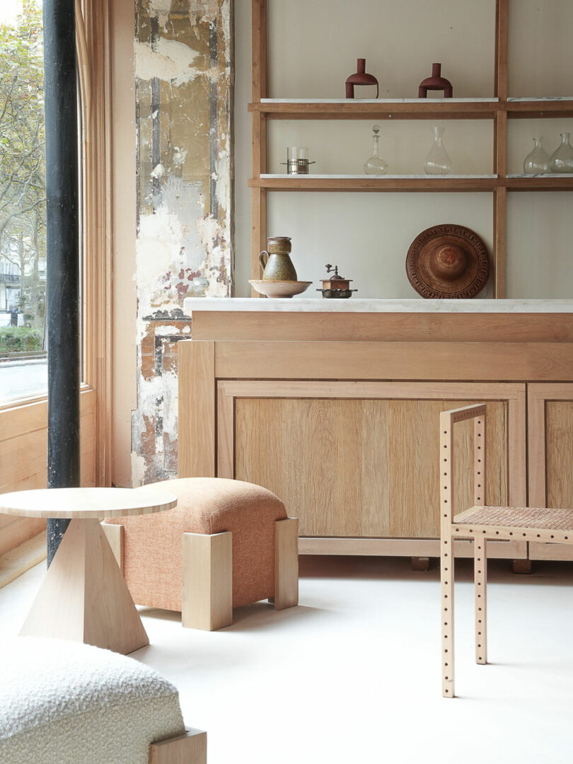 A minimalist room features wooden furniture, including a chair, table, shelves, and a cabinet. Decorative items such as vases and bowls are displayed. A large window allows natural light to pour in, enhancing the serene Policronica aesthetic.