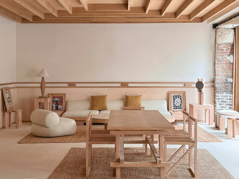 A minimalist living room featuring Policronica wooden furniture, a white sofa with two tan cushions, a unique chair, lamp, and framed art. Exposed brick and beams add rustic charm to the space.