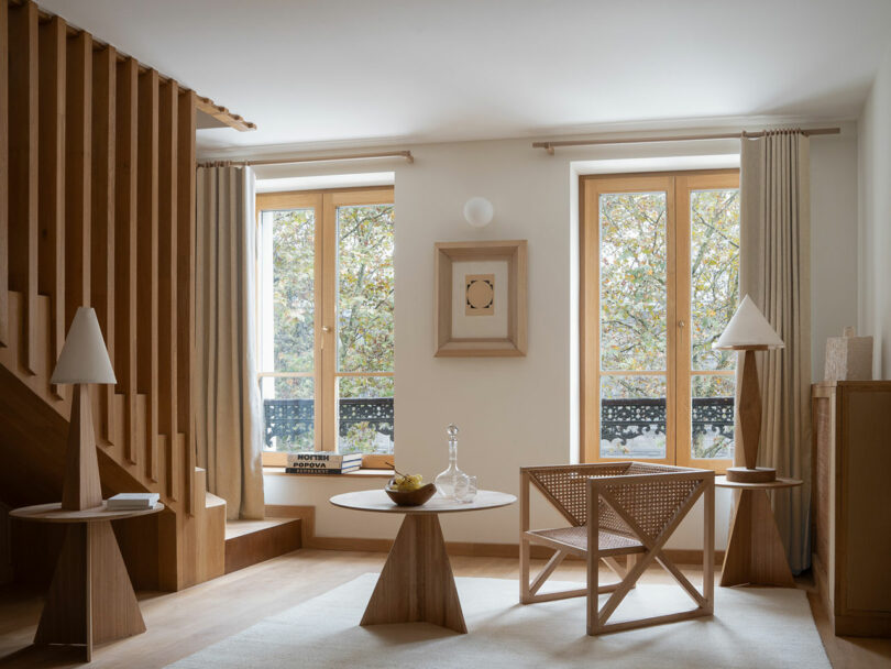 A modern living room featuring wooden furniture and two large windows with beige curtains exudes a policronica charm. A circular table, decorative lamps, and a fruit bowl add to the ambiance. On the left, stairs with wooden slats elegantly complete this stylish space.