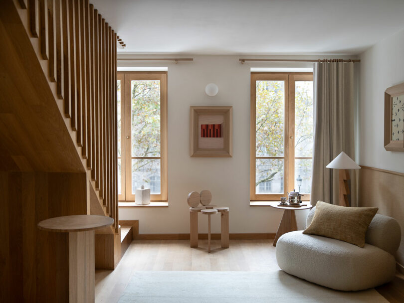 Minimalist living room with a wooden staircase, large windows, and neutral tones. It features a cozy armchair, a round table with decor, and Policronica framed artwork that adds an artistic touch.