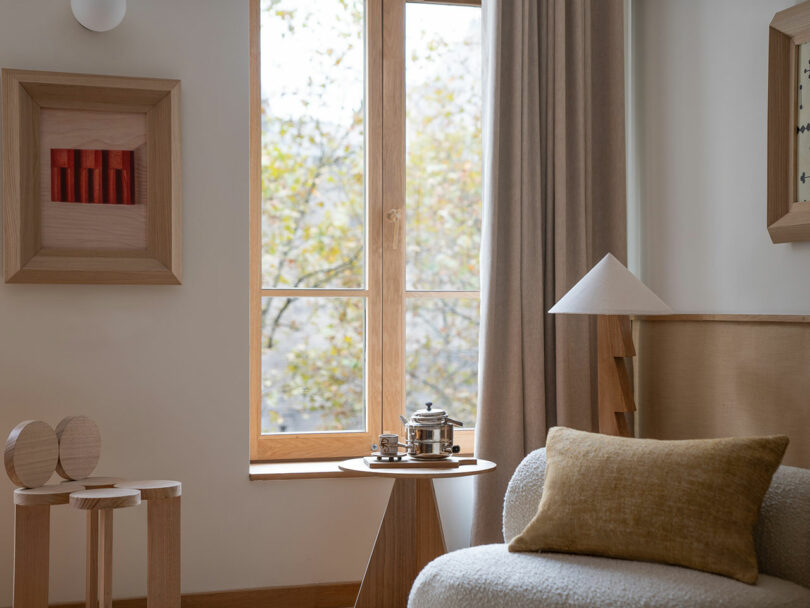 This minimalist room features wooden decor and a Policronica touch. A round side table holds a silver tea set, paired with a cream chair and mustard cushion. Framed artwork adorns the wall, while beige curtains softly filter the light through the window.