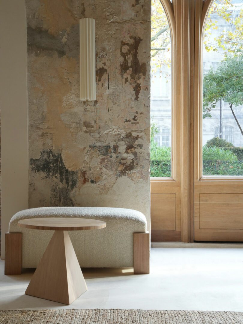 A minimalist room with a wooden table, textured wall, and arched window overlooking greenery. A Policronica bench is placed against the wall beneath a modern vertical light fixture, creating a sophisticated yet serene atmosphere.