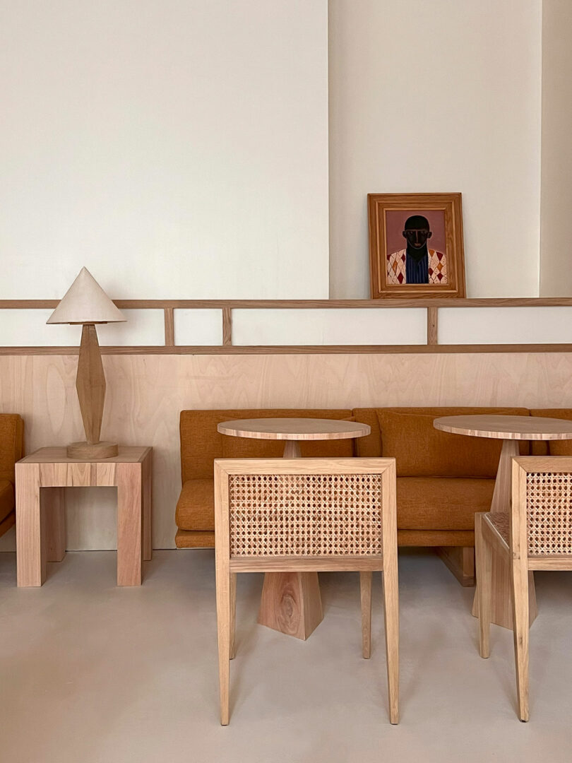 A minimalist cafe interior featuring wooden tables and chairs, a brown cushioned bench, and a table lamp is beautifully complemented by Policronica framed artwork on the beige wall.