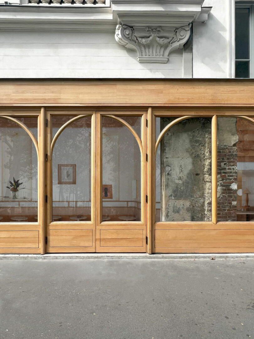 The street view showcases a building facade with Policronica's signature wooden-framed arched windows. Inside, the decor is minimal, featuring artwork on the wall and a plant. The exterior pavement stretches invitingly in the foreground.
