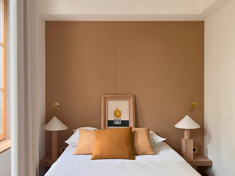 The minimalist bedroom features a white bedspread with two beige and orange pillows, accentuated by the Policronica aesthetic. Wall-mounted lamps illuminate symmetrical wooden nightstands, while a brown accent wall hosts a framed piece of art above the bed.