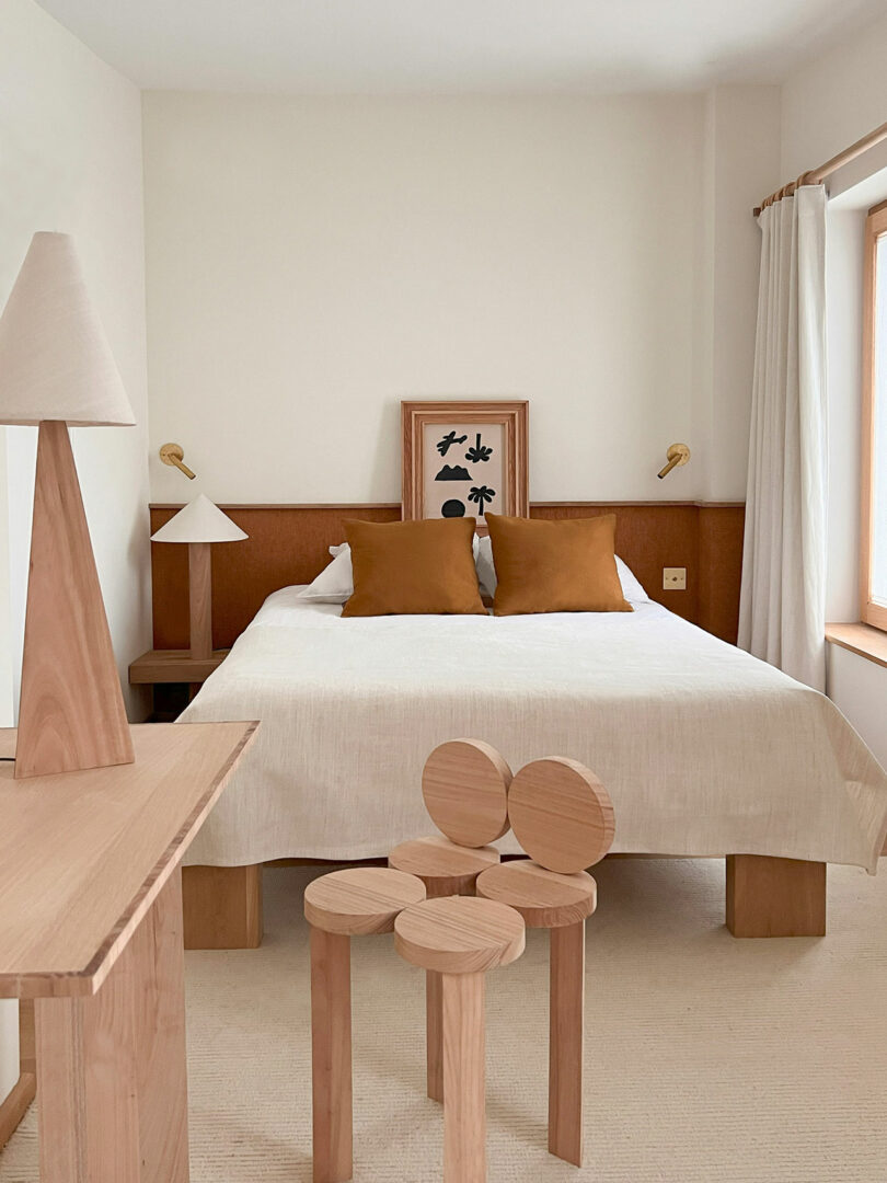 The minimalist bedroom features a double bed with white bedding and brown pillows, complemented by two wooden lamps on side tables. A wooden chair sits in front, while Policronica reflects in the framed artwork above the bed.