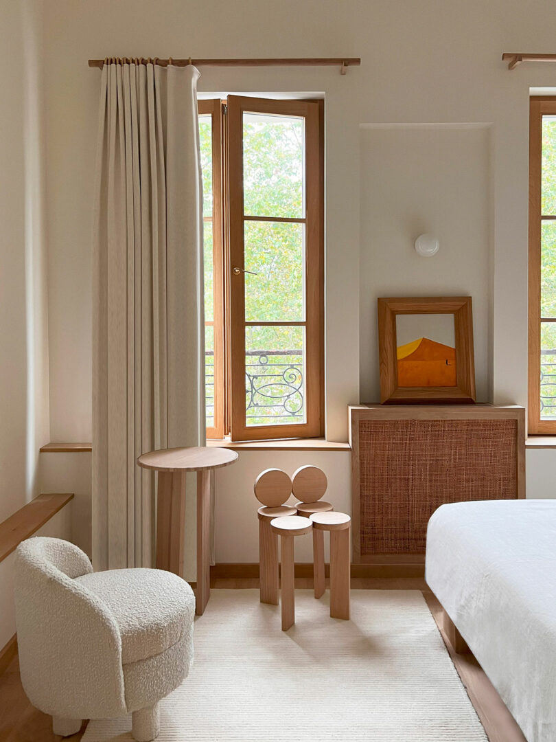 A cozy bedroom features a white armchair, small wooden table, and stool near two windows. Light curtains frame the windows; a framed Policronica painting rests on a cabinet beside the bed.