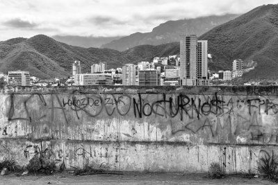 A black-and-white image of the skyline of Monterrey, Mexico with rolling hills in the background. In the foreground is a graffitied wall. 